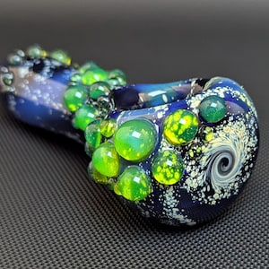May include: A blue and white glass pipe with a swirling design and green glass accents. The pipe is decorated with small, round, green and yellow glass beads.