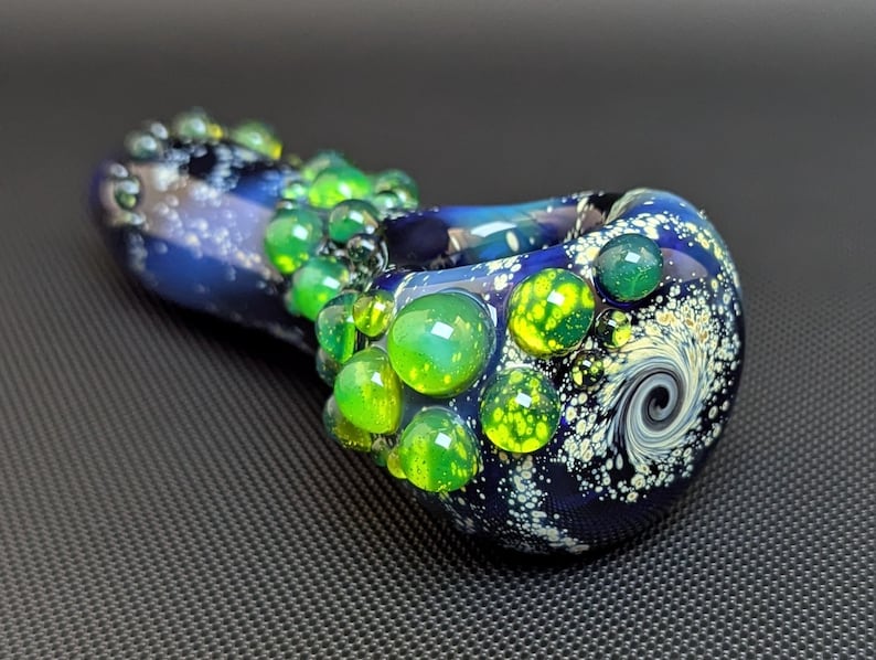 May include: A blue and white glass pipe with a swirling design and green glass accents. The pipe is decorated with small, round, green and yellow glass beads.