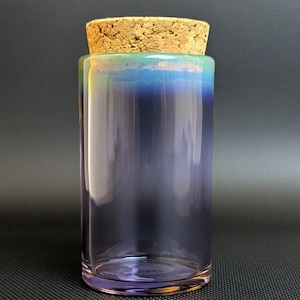 Pode incluir: Um frasco de vidro cilíndrico com uma rolha de cortiça. O frasco tem um gradiente de cores, passando do lilás claro na base a um tom azul-esverdeado na borda. A rolha de cortiça é de cor castanho claro.