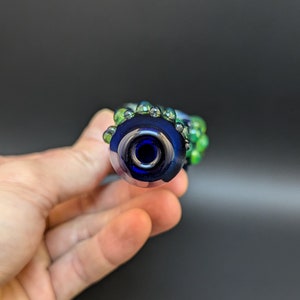 May include: A blue glass pipe with green glass accents. The pipe has a round bowl and a small opening for smoking.