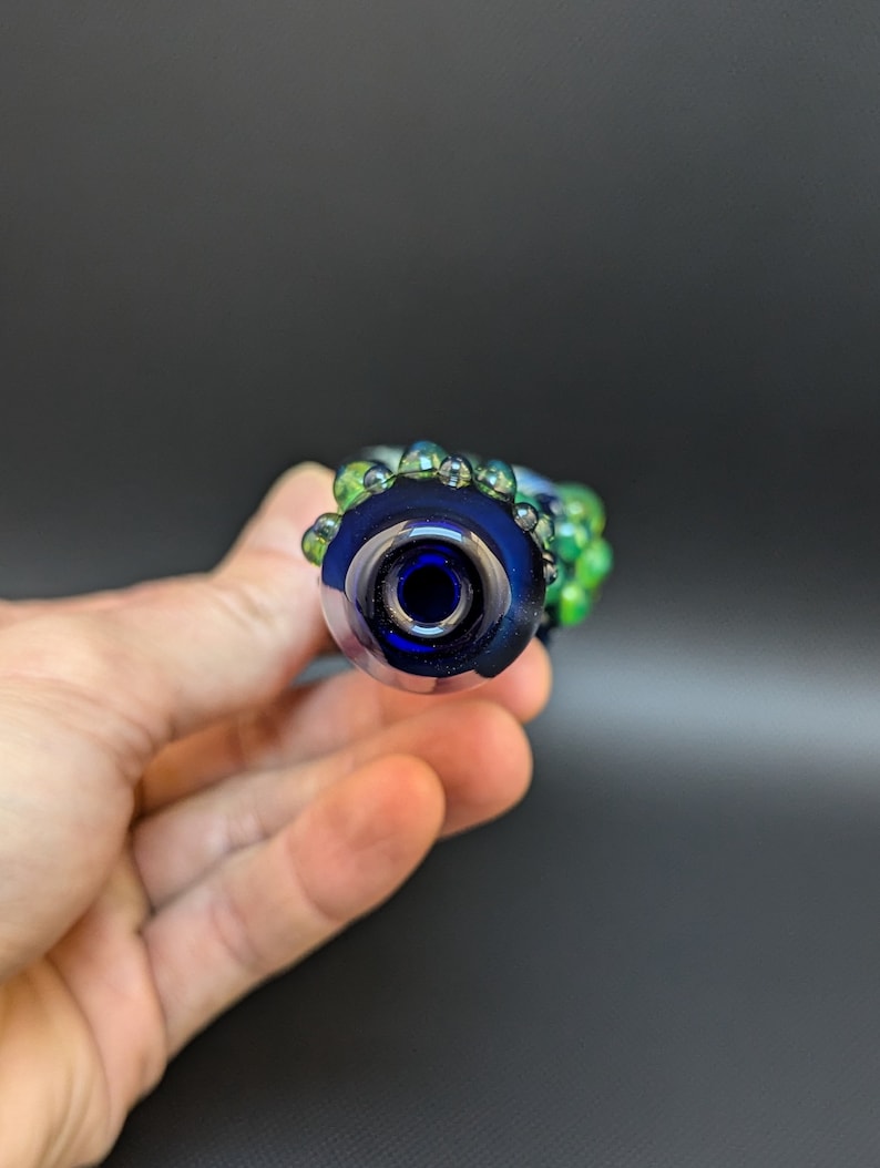 May include: A blue glass pipe with green glass accents. The pipe has a round bowl and a small opening for smoking.