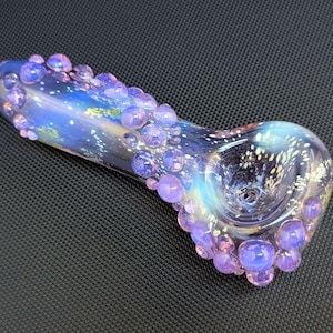 May include: A hand-blown glass pipe with a galaxy-inspired design. The pipe features a blue and purple color scheme with white speckles, and is adorned with numerous purple glass bubbles. The bowl has a circular opening.