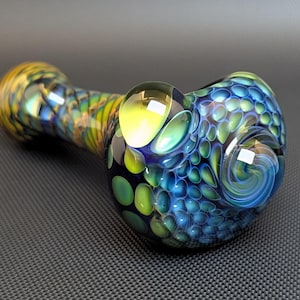 May include: A colorful glass pipe with a rounded bowl and a long stem. The pipe features a blend of blue, green, yellow, and gold hues. The bowl has a textured pattern, and the stem has a swirling design.