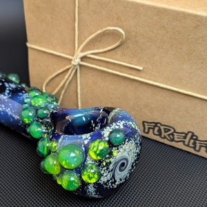 May include: A glass pipe with a galaxy design and green accents. The pipe is made of glass and has a round bowl. The pipe is sitting on a black surface. The text "Firelife" is visible on the box.