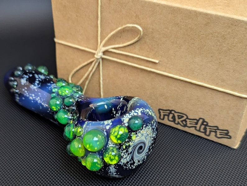 May include: A glass pipe with a galaxy design and green accents. The pipe is made of glass and has a round bowl. The pipe is sitting on a black surface. The text "Firelife" is visible on the box.