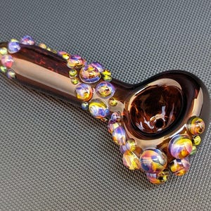 May include: A brown glass pipe with a bowl and a long stem. The stem is decorated with iridescent glass beads in shades of purple, yellow, and blue.