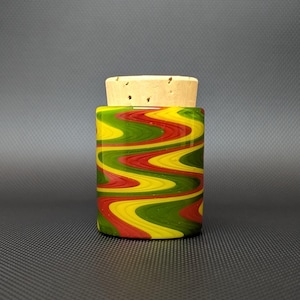 May include: A cylindrical glass jar with a cork stopper. The jar is decorated with a wavy pattern of red, yellow, and green. The cork stopper is a light brown color. The jar is on a dark gray surface.