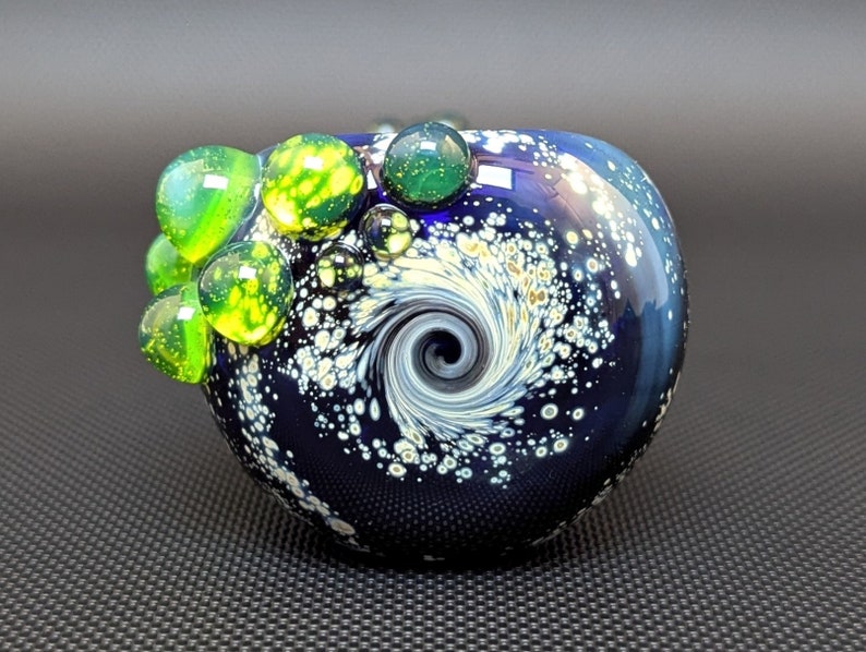 May include: A blue glass pipe with a swirling white and gold design. The pipe has a small opening on the side and is decorated with green and yellow glass beads.