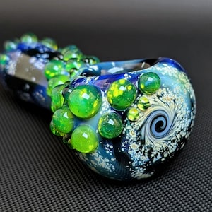 May include: A glass pipe with a blue, green, and white galaxy design. The pipe has a swirl pattern and is decorated with green glass bubbles.