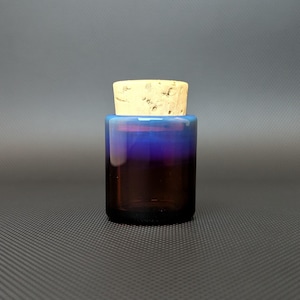 Pode incluir: Um pequeno frasco cilíndrico de vidro com uma rolha de cortiça. O frasco faz a transição de uma base castanha escura para uma borda azul vibrante. A cortiça é de cor castanho claro. O frasco é provavelmente usado para armazenar pequenos itens ou líquidos.