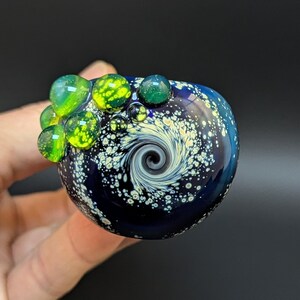 May include: A small, round, blue glass orb with a swirling white and black design. The orb is decorated with green and yellow glass bubbles.