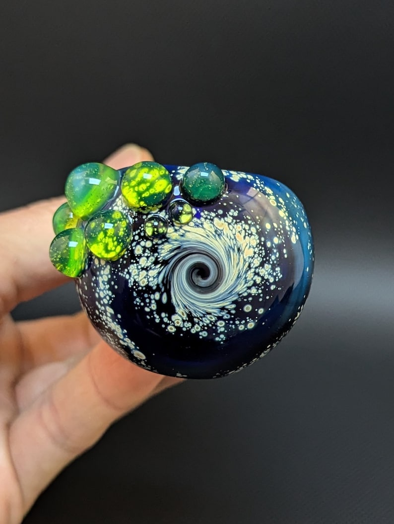 May include: A small, round, blue glass orb with a swirling white and black design. The orb is decorated with green and yellow glass bubbles.