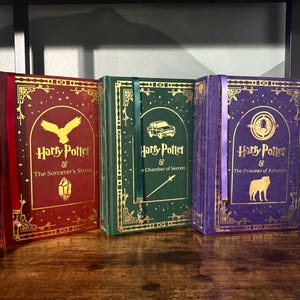 May include: Three hardcover books from the Harry Potter series are displayed on a wooden shelf. The books are in red, green, and purple with gold lettering and illustrations. Each book has a ribbon bookmark.