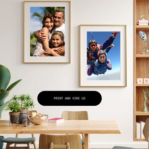 May include: Two framed photographs hang on a white wall. One features a family portrait, the other shows three people skydiving. A black oval below the photos reads "PRINT AND VIBE US". A wooden table with plants and fruit is in the foreground.