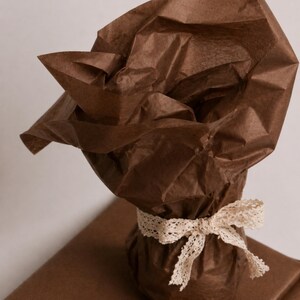 May include: A wrapped gift with brown tissue paper and a cream-colored lace ribbon tied in a bow. The gift is on top of a brown wrapping paper. The tissue paper is crumpled and adds texture to the presentation.