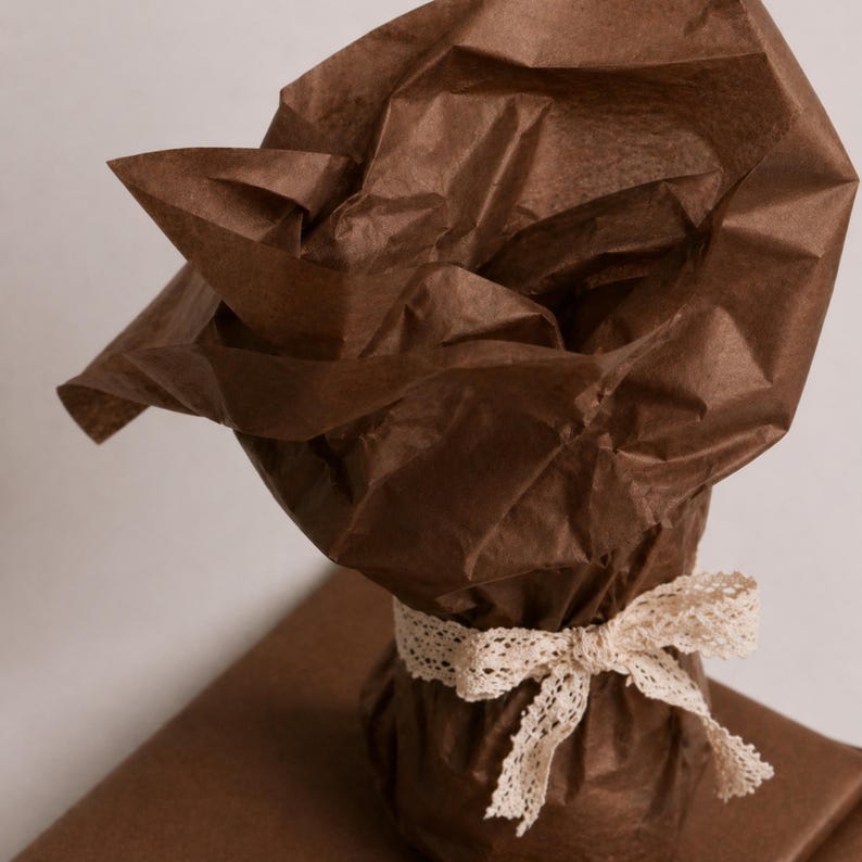 May include: A wrapped gift with brown tissue paper and a cream-colored lace ribbon tied in a bow. The gift is on top of a brown wrapping paper. The tissue paper is crumpled and adds texture to the presentation.