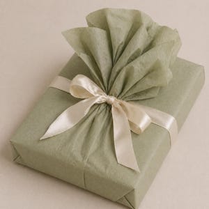 May include: A wrapped gift in sage green paper, adorned with a cream-colored satin ribbon tied in a bow. The top of the package is embellished with a fan of matching tissue paper, creating a decorative presentation.