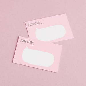 May include: Two pink notecards with a white speech bubble design and the text "A note to say...". The cards are arranged diagonally against a pink background, creating a simple and clean aesthetic.
