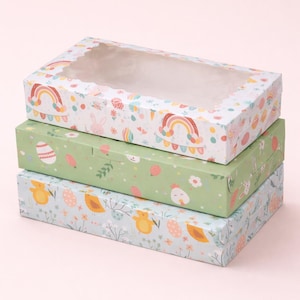 May include: Three stacked rectangular gift boxes with Easter patterns. The top box has a clear window and a white background with rainbows, bunnies, and flowers. The middle box is green with Easter eggs and leaves. The bottom box is light blue with chicks and eggs.