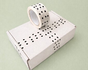 May include: A roll of white tape with black polka dots sits atop a white cardboard box. The box is sealed with matching polka dot tape. The background is a solid light green.