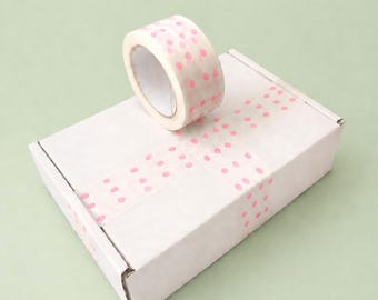 May include: A roll of decorative tape with pink polka dots sits atop a white cardboard box. The tape is also used to seal the box, creating a cohesive design. The background is a solid light green.