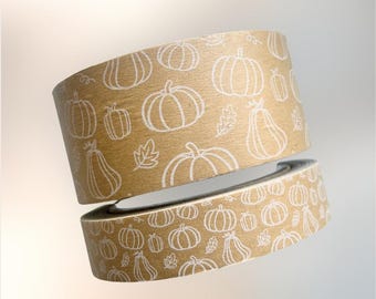 May include: Two rolls of decorative tape with a kraft paper background. The tape is patterned with white line drawings of pumpkins and leaves. The rolls are slightly separated, showing the design on the edges.