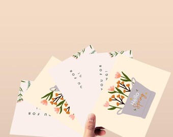 May include: A hand holding a collection of greeting cards with floral designs. The cards feature illustrations of flowers, leaves, and the phrase "Hello Spring". The color palette includes soft pastels and earth tones.