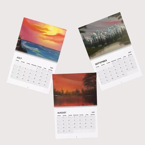 May include: Three wall calendars for the year 2027, each featuring a different landscape painting at the top. The July calendar shows a sunset over the ocean, August displays a red-toned forest lake, and September depicts snow-capped mountains.