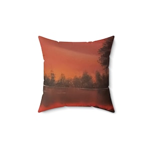 以下が含まれることがあります： 夕焼けの風景画が描かれた正方形のクッション。燃えるようなオレンジ色の空、暗い並木、反射する水面が描かれています。クッションは柔らかく、質感があり、どんな部屋にもアート感を加えるのに最適です。