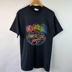 May include: Black t-shirt with a vibrant graphic celebrating the 30th anniversary of Universal Studios Hollywood. The design includes fireworks in pink, orange, yellow, green, and blue. The text reads "UNIVERSAL STUDIOS Hollywood 30th ANNIVERSARY 1964".