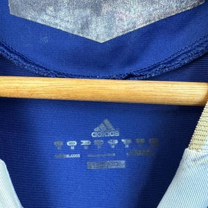 May include: Close-up of a blue Adidas athletic jersey. The jersey has a silver panel at the top and a white and light blue collar. The Adidas logo and size tags are visible.