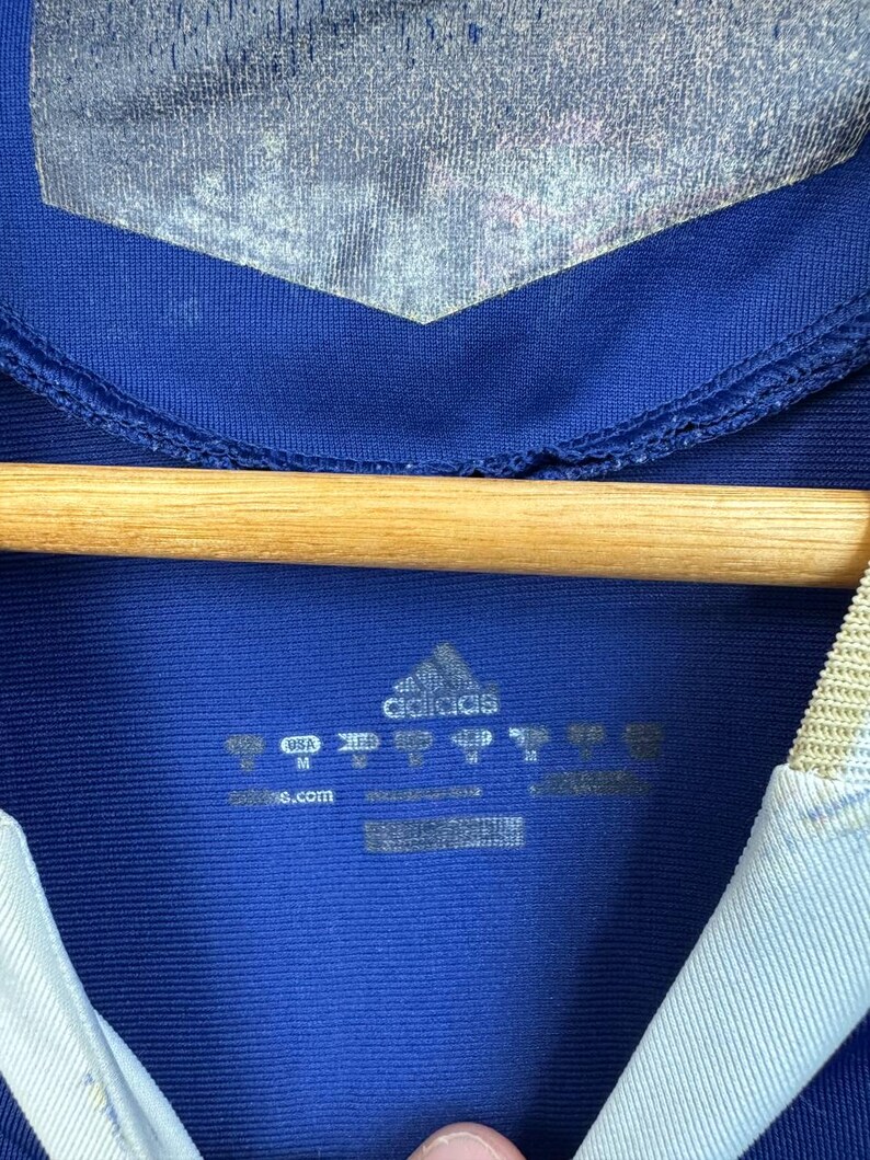May include: Close-up of a blue Adidas athletic jersey. The jersey has a silver panel at the top and a white and light blue collar. The Adidas logo and size tags are visible.