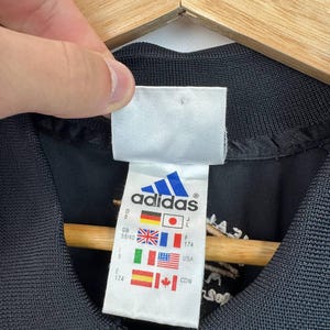 May include: Close-up of a black Adidas jacket with a white tag. The tag features the Adidas logo and flags of several countries, including Germany, Japan, the UK, France, Italy, the USA, and Canada. The jacket has a zipper and a ribbed collar.