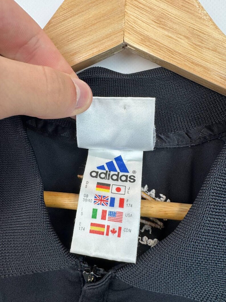 May include: Close-up of a black Adidas jacket with a white tag. The tag features the Adidas logo and flags of several countries, including Germany, Japan, the UK, France, Italy, the USA, and Canada. The jacket has a zipper and a ribbed collar.