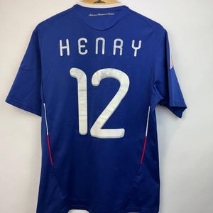 May include: A blue soccer jersey with the name "HENRY" and the number "12" in white lettering on the back. The jersey has short sleeves and a small French flag detail. The jersey is hanging on a wooden hanger.