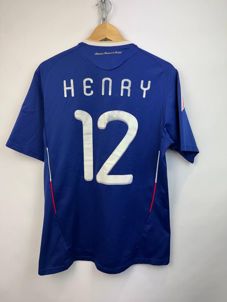 May include: A blue soccer jersey with the name "HENRY" and the number "12" in white lettering on the back. The jersey has short sleeves and a small French flag detail. The jersey is hanging on a wooden hanger.