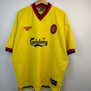 May include: A yellow Liverpool soccer jersey with a V-neck collar and short sleeves. The jersey features the Carlsberg logo, the Reebok logo, and a team crest. The collar and sleeve cuffs have red and black trim. The jersey is hanging on a metal hanger.