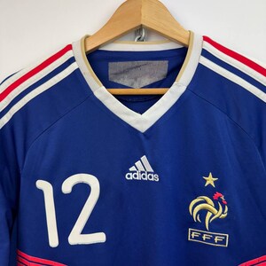 May include: A blue soccer jersey with white and red stripes on the shoulders. The number 12 is on the front, along with the Adidas logo and a French emblem. The jersey is hanging on a wooden hanger.
