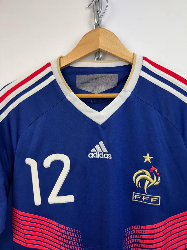 May include: A blue soccer jersey with white and red stripes on the shoulders. The number 12 is on the front, along with the Adidas logo and a French emblem. The jersey is hanging on a wooden hanger.