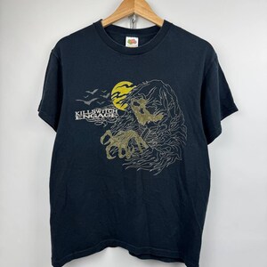 May include: A black short-sleeved t-shirt featuring a graphic print of a skeletal figure with a clawed hand, a yellow moon, and a flock of birds. The text "KILLSWITCH ENGAGE" is printed in white on the left side of the chest area.