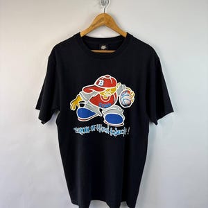 Könnte beinhalten: Schwarzes T-Shirt mit einer Cartoon-Illustration eines Basketballspielers mit roter Mütze und blauen Schuhen. Der Text "School of Hard Knock" ist unter der Illustration aufgedruckt.