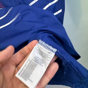 May include: Close-up of a blue athletic shirt with white and red accents. The shirt has a tag with text in multiple languages. The shirt is made of 100% polyester.