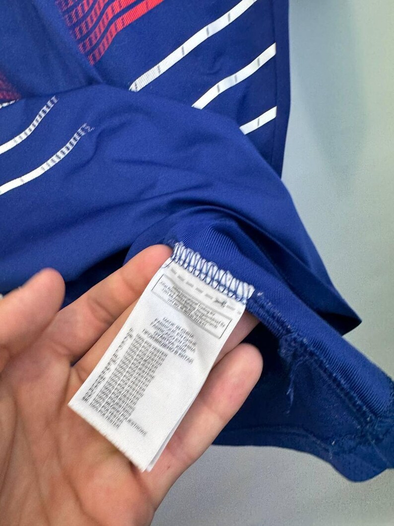 May include: Close-up of a blue athletic shirt with white and red accents. The shirt has a tag with text in multiple languages. The shirt is made of 100% polyester.