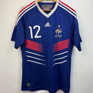 May include: A blue soccer jersey with white and red accents, featuring the number 12 in white. The Adidas logo and the French Football Federation emblem are visible. The jersey has a V-neck and is hanging on a wooden hanger.