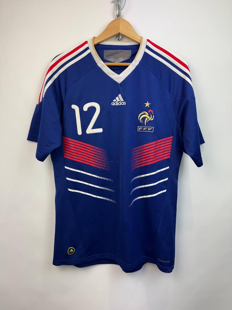 May include: A blue soccer jersey with white and red accents, featuring the number 12 in white. The Adidas logo and the French Football Federation emblem are visible. The jersey has a V-neck and is hanging on a wooden hanger.