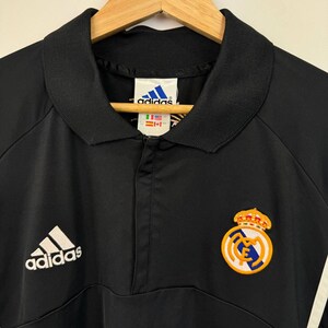 May include: Black Adidas Real Madrid soccer jacket with a collared neckline and a half-zip closure. Features the Adidas logo on the left chest and the Real Madrid crest on the right. Includes white stripes on the right sleeve.