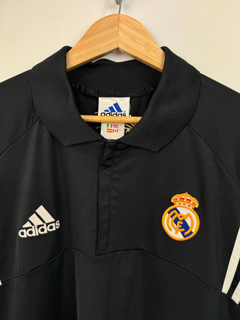 May include: Black Adidas Real Madrid soccer jacket with a collared neckline and a half-zip closure. Features the Adidas logo on the left chest and the Real Madrid crest on the right. Includes white stripes on the right sleeve.