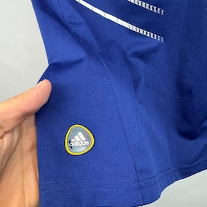 May include: Royal blue athletic shirt with white stripes and a logo patch. The shirt features a crew neck and short sleeves. The Adidas logo is visible on the lower left side of the shirt.
