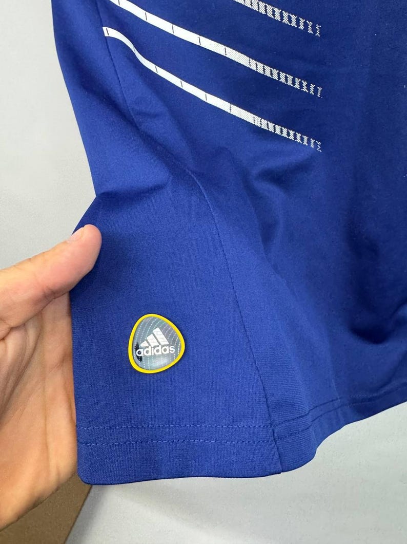 May include: Royal blue athletic shirt with white stripes and a logo patch. The shirt features a crew neck and short sleeves. The Adidas logo is visible on the lower left side of the shirt.