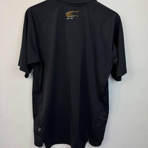 May include: Black polo shirt with short sleeves, hanging on a wooden hanger. The back of the shirt features a small gold and white logo with the text "REAL MADRID" and a smaller line of text below it. The shirt is made of a smooth, possibly athletic material.